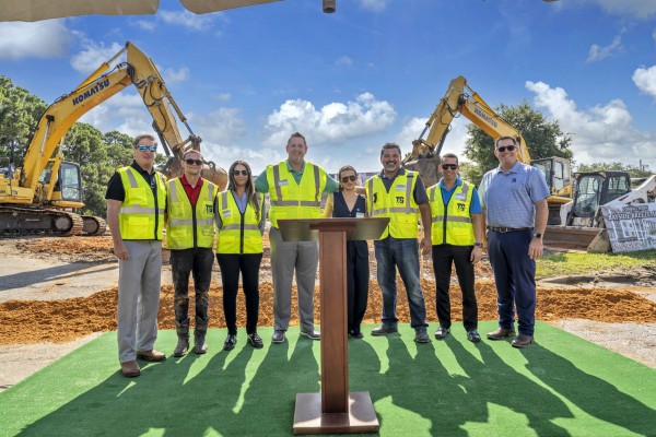 Lineup of eight people at the groundbreaking ceremony for Bayside Breeze. Podium in front of the people. Construction equipment in the background.