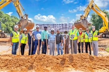 Lineup of people leaning on shovels in the dirt at the groundbreaking ceremony for Bayside Breeze. Large pile of dirt front of the people. Construction equipment in the background.